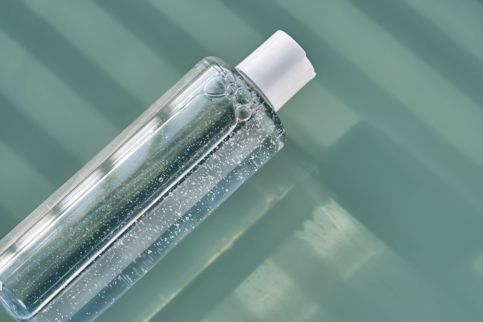 Bottle of facial toner or cleanser on a green background with stripes shadows.